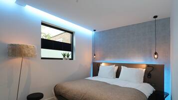 Een moderne slaapkamer met een groot bed, een klein krukje, een nachtkastje en een raam met jaloezieën.