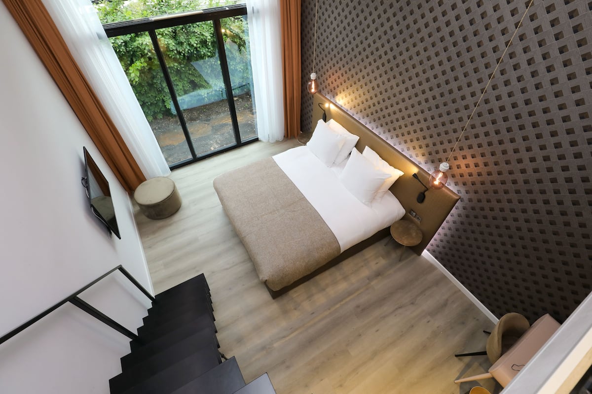 A modern hotel room with a large bed, a small round table, a sofa, and a view of greenery through a sliding door.