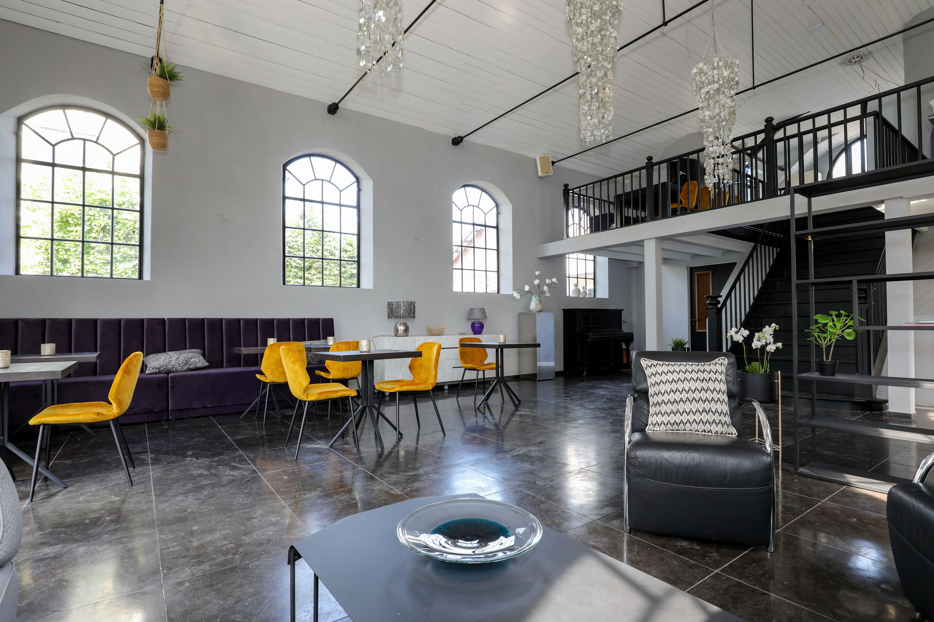 A spacious living area with a high ceiling, large windows, a dining area with yellow chairs, and a staircase leading to an upper level.
