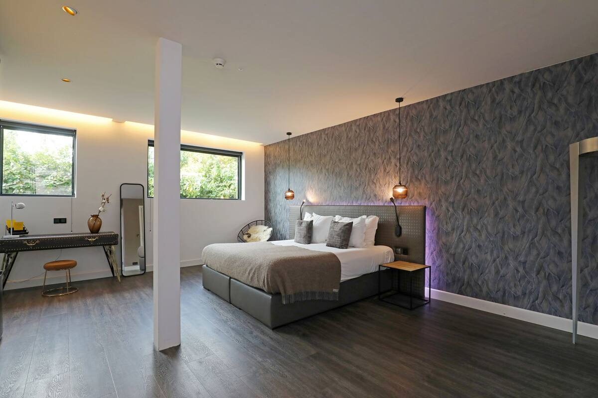 A modern bedroom with a large bed, a desk, and a chair. There are windows providing natural light and a view of greenery outside.