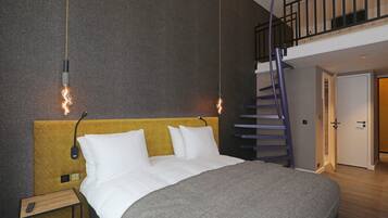 A modern hotel room with a large bed, a wooden nightstand, a wall-mounted light fixture, and a staircase to an upper level.