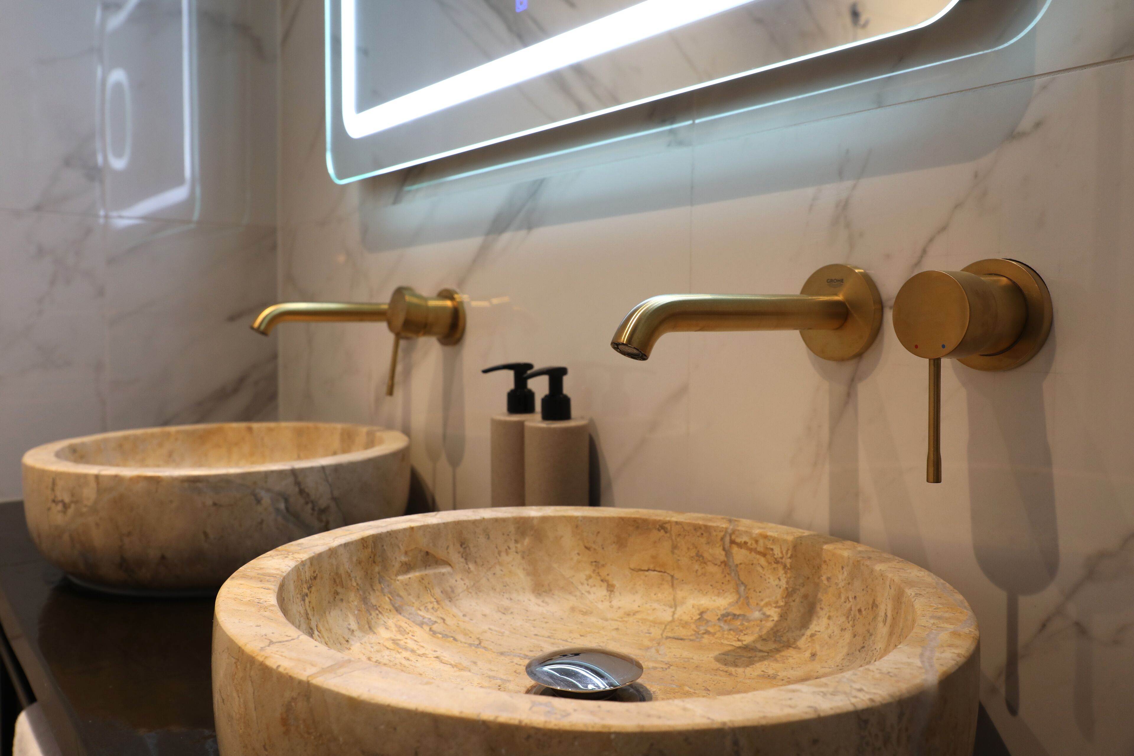 A bathroom with two marble sinks, gold faucets, and a modern mirror with LED lighting.