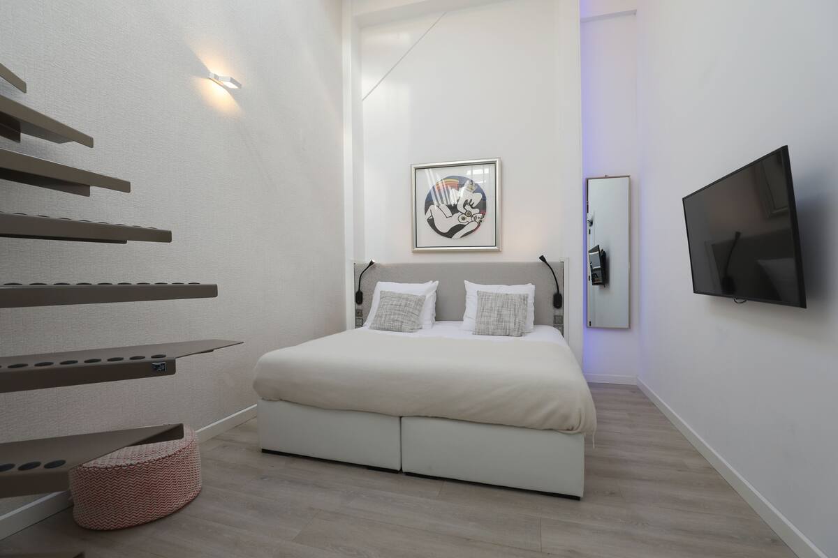 A modern bedroom with a bed, a flat-screen TV, a framed artwork, and a staircase.