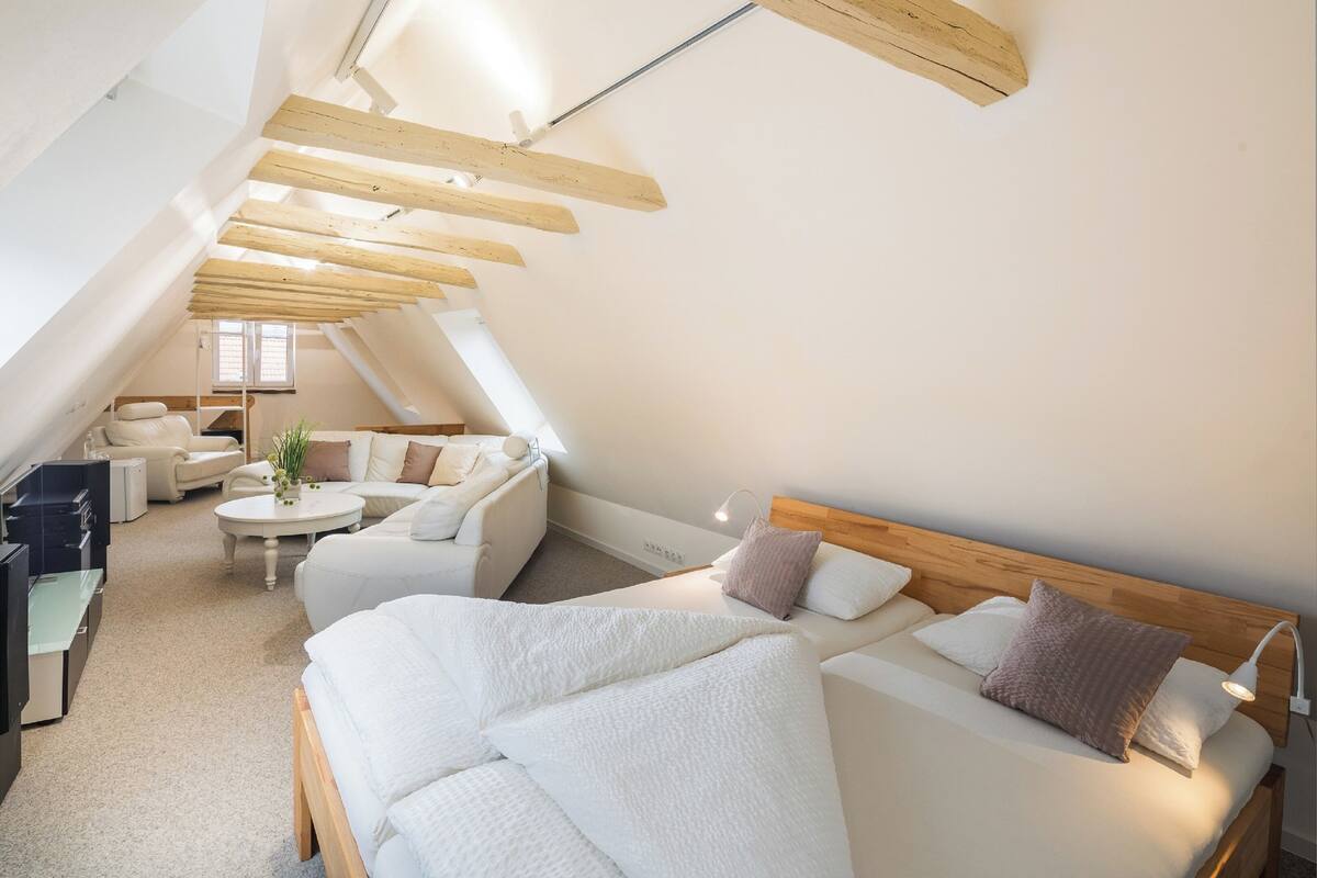 A modern, well-lit attic bedroom with a sloped ceiling, a large bed, a sofa, a small table, and a desk.