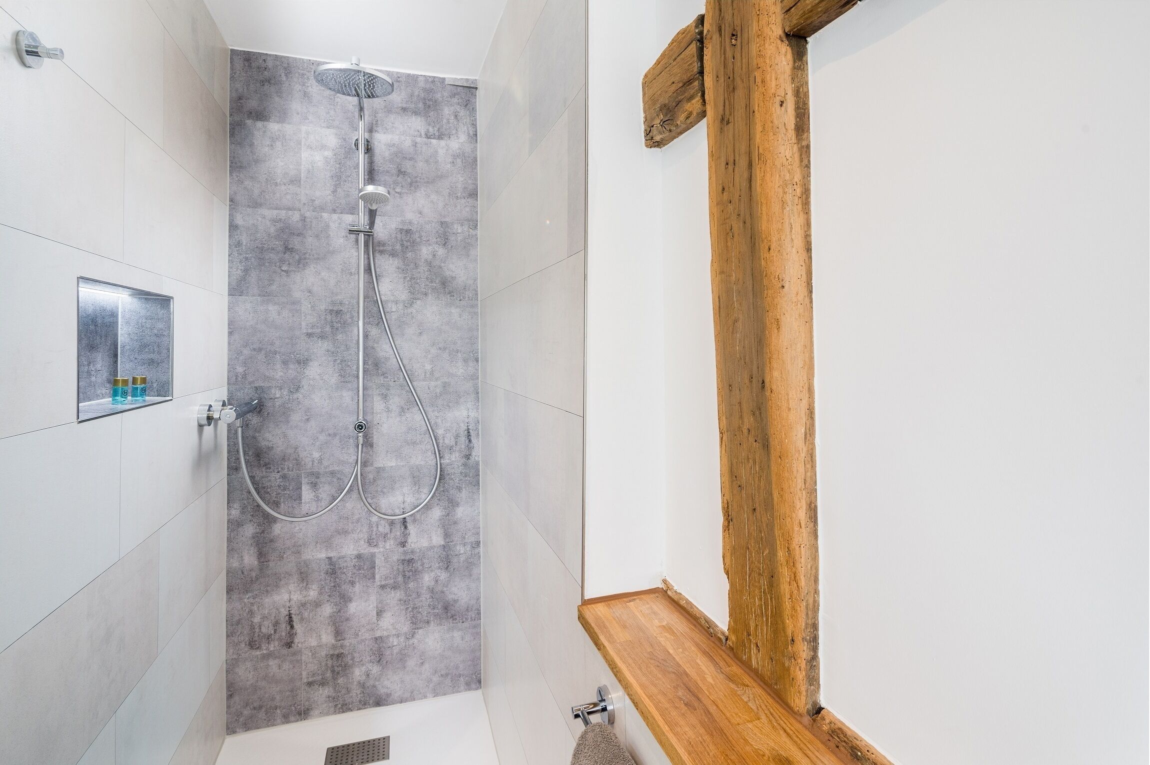 A shower with a grey textured wall, a wooden bench, and a metal faucet.