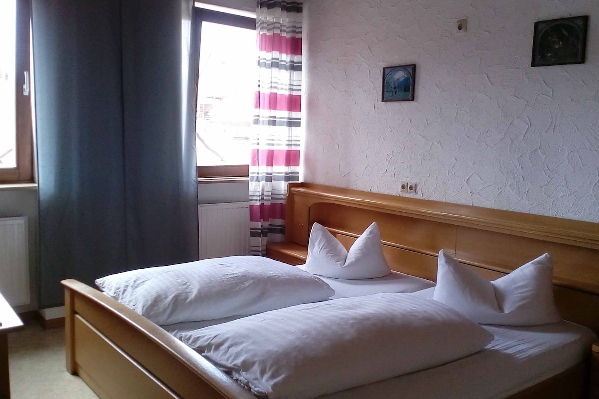 A bedroom with a wooden bed, two pillows, a small bedside table, a window with curtains, and a wall-mounted picture.