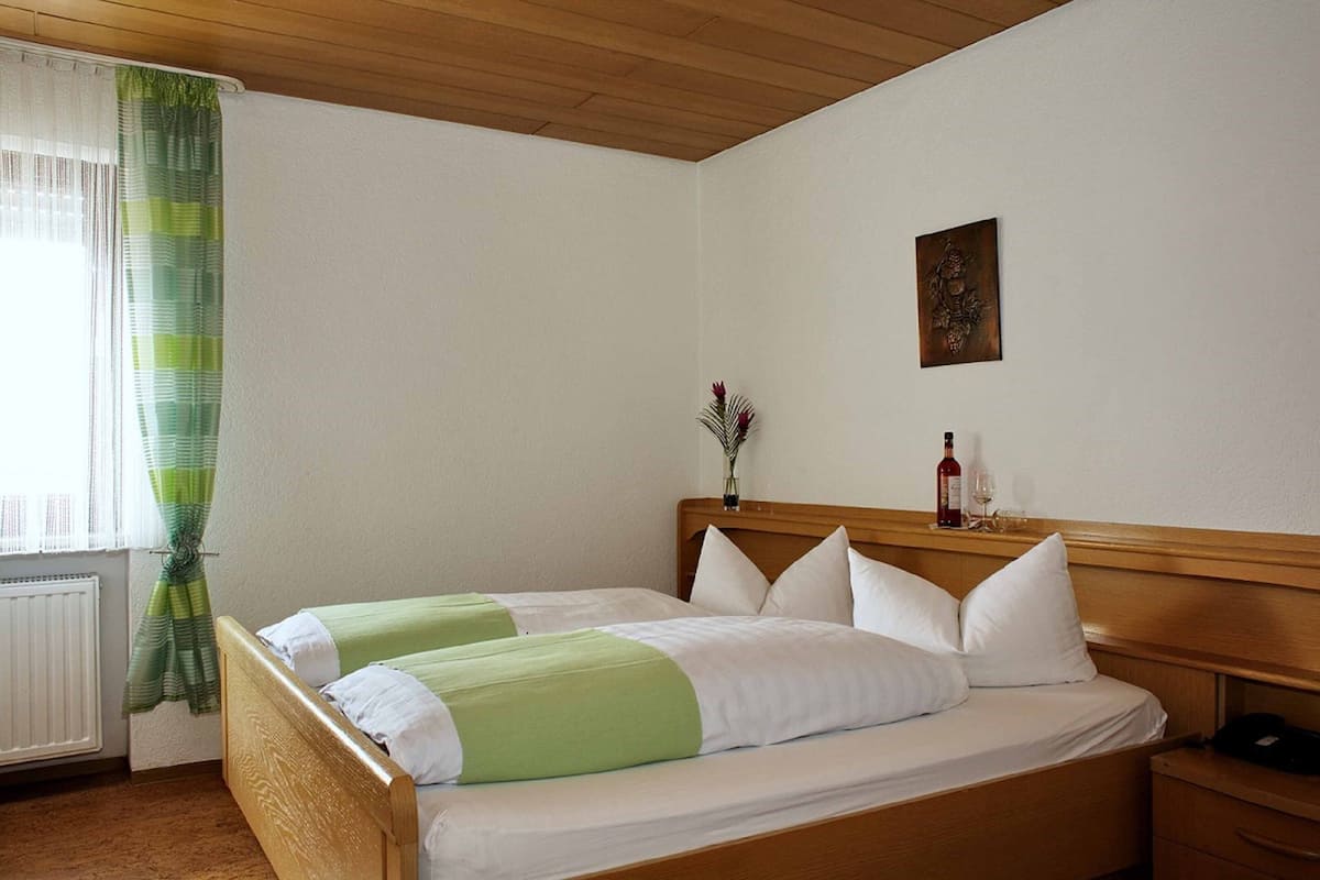A hotel room with two single beds, a wooden headboard, a radiator, a window with green curtains, a wall-mounted artwork, and a bedside table with a bottle and glass.