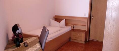 A small, single-bed room with a wooden headboard, a bedside table with a vase of flowers, and a chair.