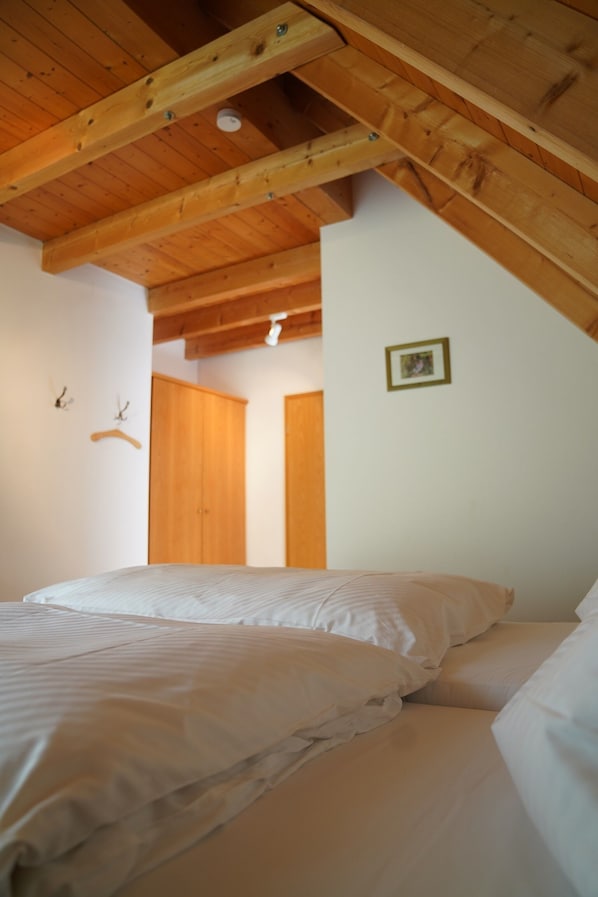 Un dormitorio con techo inclinado, vigas de madera, una cama con ropa blanca, un armario y un cuadro enmarcado en la pared.