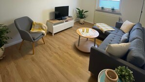 A modern living room with a grey sofa, a grey armchair, a small round coffee table, a TV on a white cabinet, and a potted plant. - Haus Landruhe, Deutschland (Neuharlingersiel)