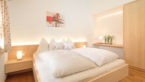 A modern bedroom with a wooden headboard, a bedside table with a lamp, a nightstand, and a wall-mounted shelf with a plant. - Vacation apartment 2 rooms - Felder Apartments (Schoppernau)