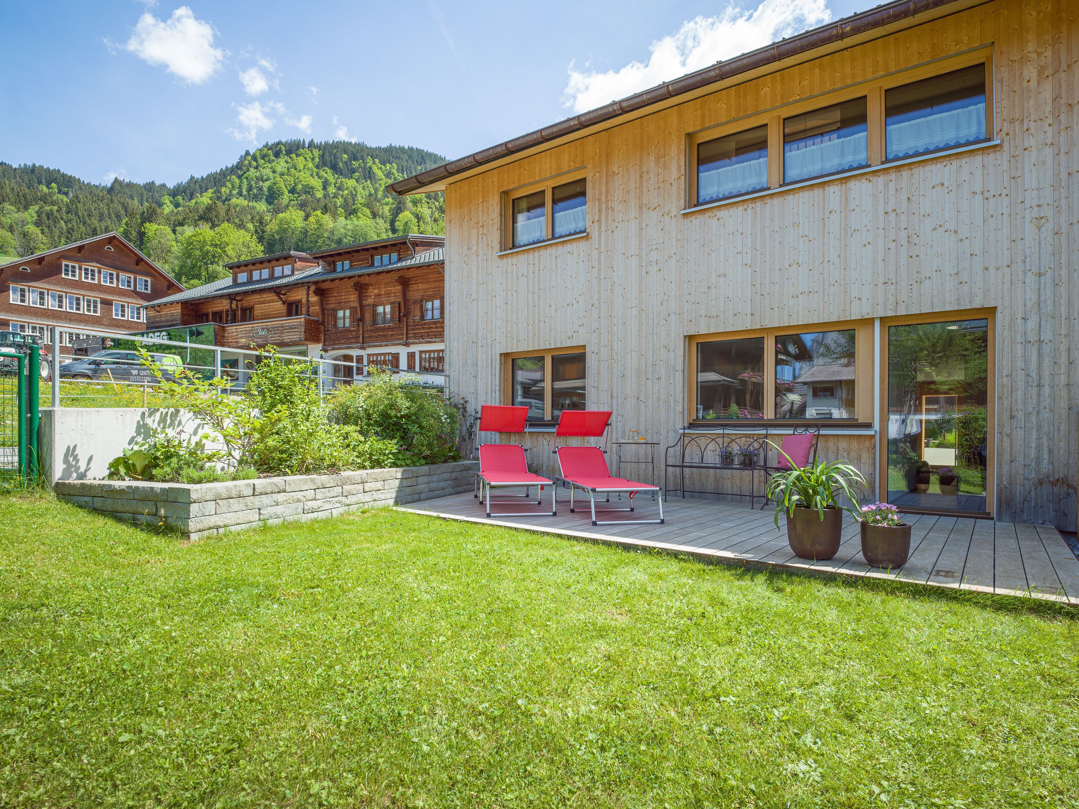 A modern wooden building with a deck, red lounge chairs, and a garden area.