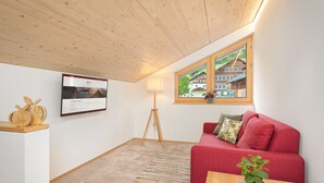 A room with a red sofa, a wooden ceiling, a window with a view, a lamp, and a framed picture on the wall. - Vacation apartment 2 rooms - Felder Apartments (Schoppernau)