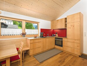 A compact kitchen with wooden cabinets, a red countertop, a built-in oven, and a window overlooking a house. - 2-room vacation apartment - Felder Apartments (Schoppernau)