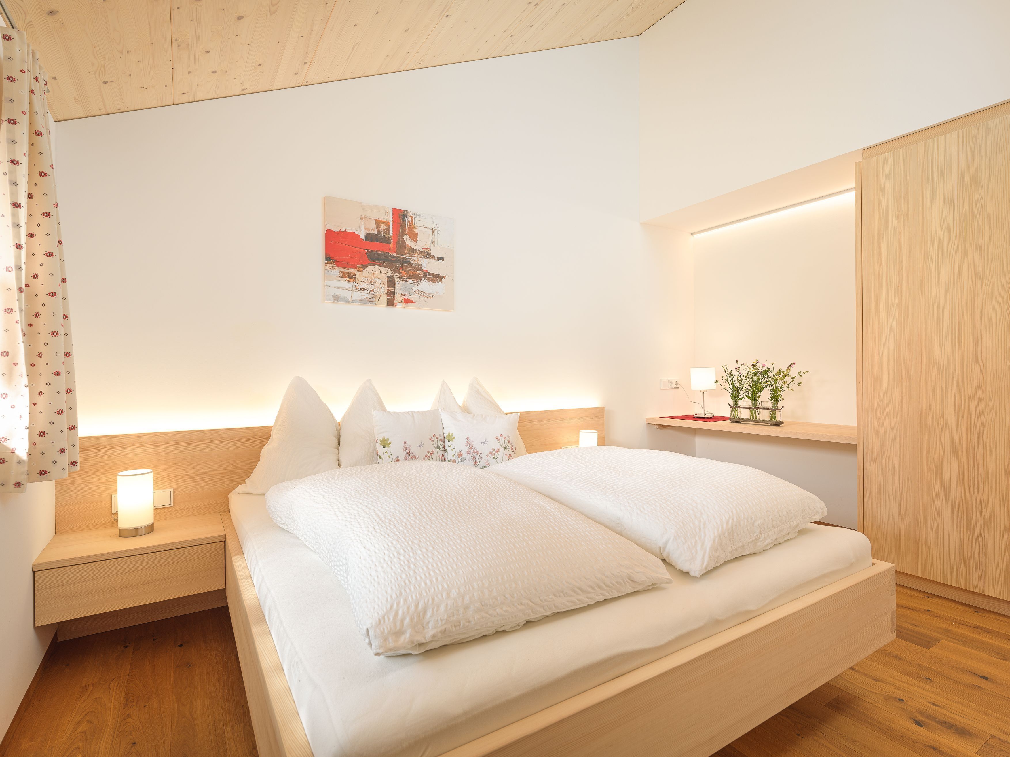 A modern bedroom with a wooden headboard, a bedside table with a lamp, a nightstand, and a wall-mounted shelf with a plant.