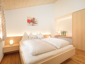 A modern bedroom with a wooden headboard, a bedside table with a lamp, a nightstand, and a wall-mounted shelf with a plant. - 2-room vacation apartment - Felder Apartments (Schoppernau)