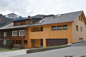 A two-story wooden house with a garage, a balcony, and a mountain backdrop. - 2-room vacation apartment - Felder Apartments (Schoppernau)