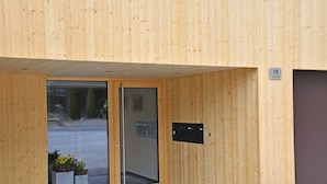 Ingresso di un edificio in legno con porta in vetro, una targa con il numero 10 e due fioriere con fiori gialli.