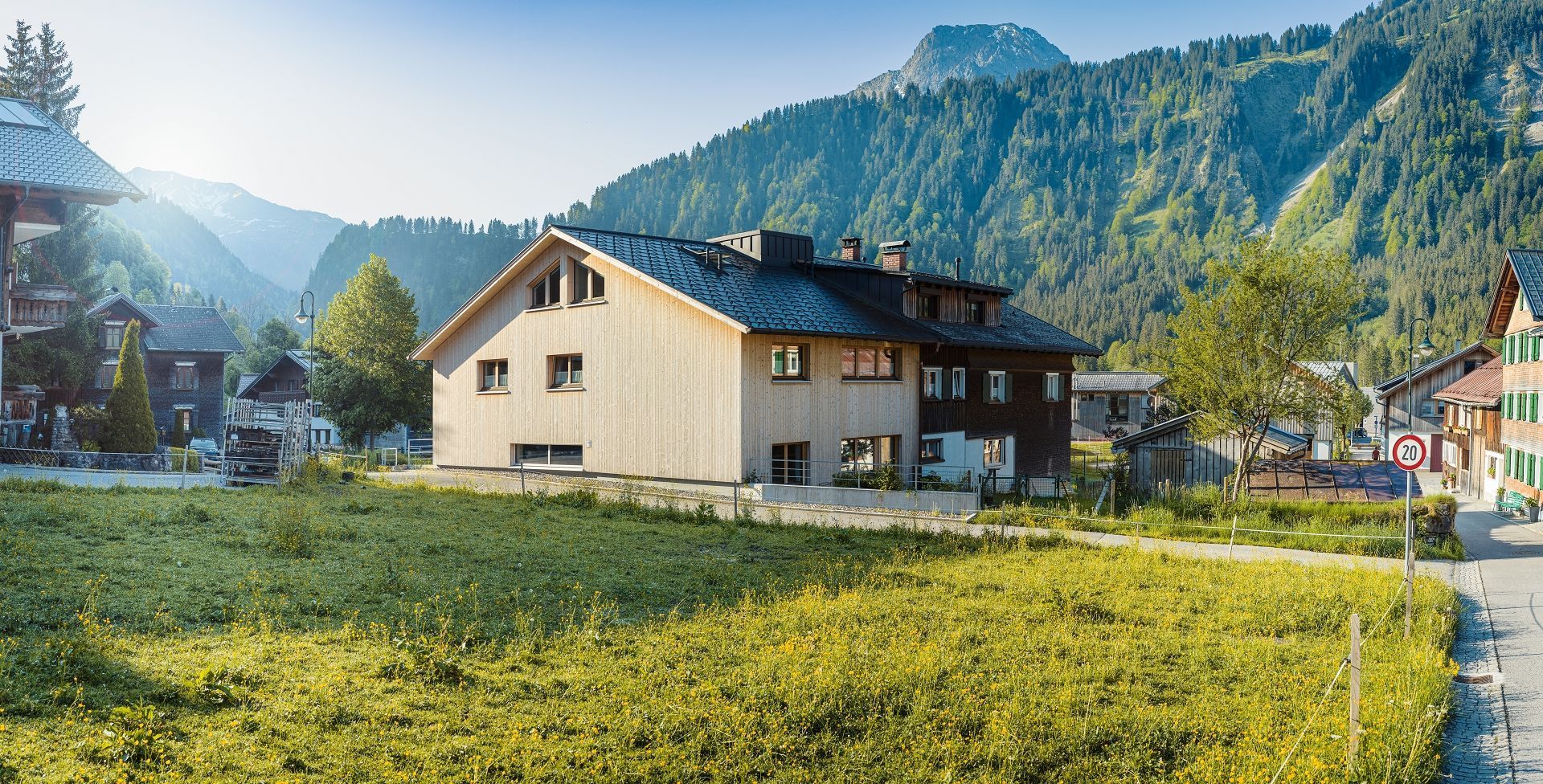 A two-story building with a sloped roof, surrounded by a grassy field and a mountain backdrop.
