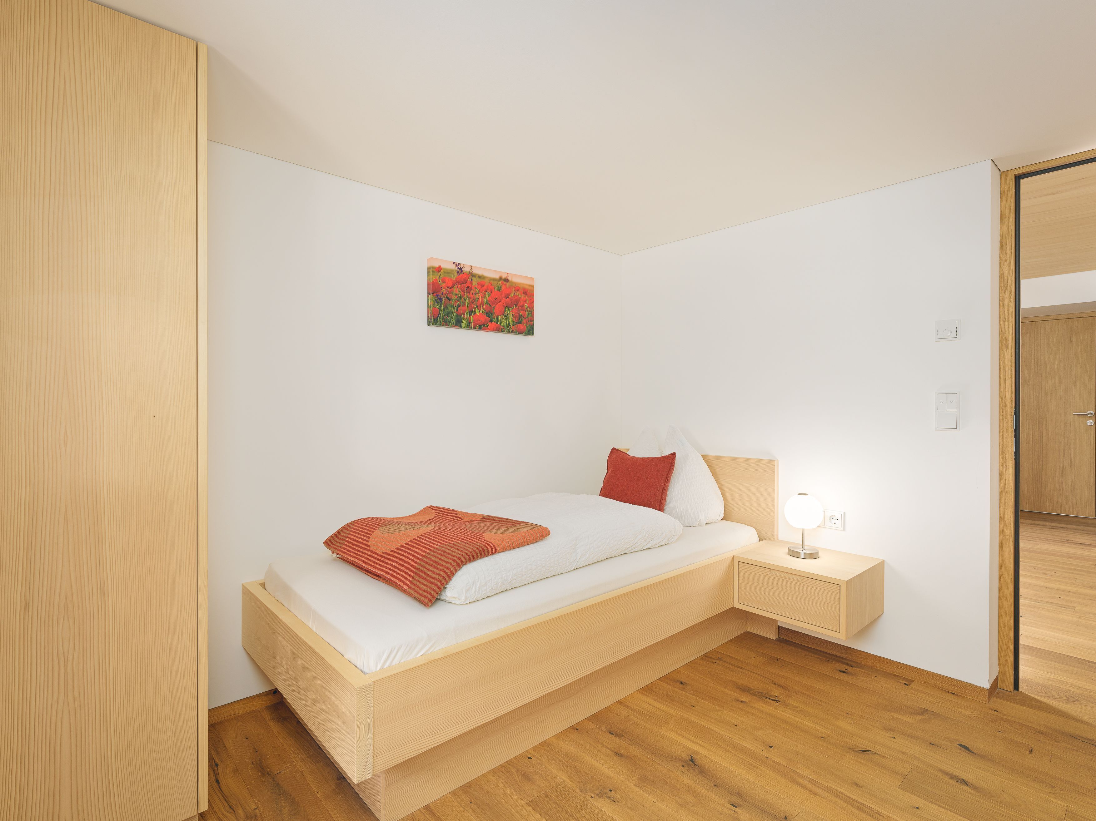 Una cama individual con estructura de madera, una mesita de noche con lámpara y un pequeño cajón junto a la cama.