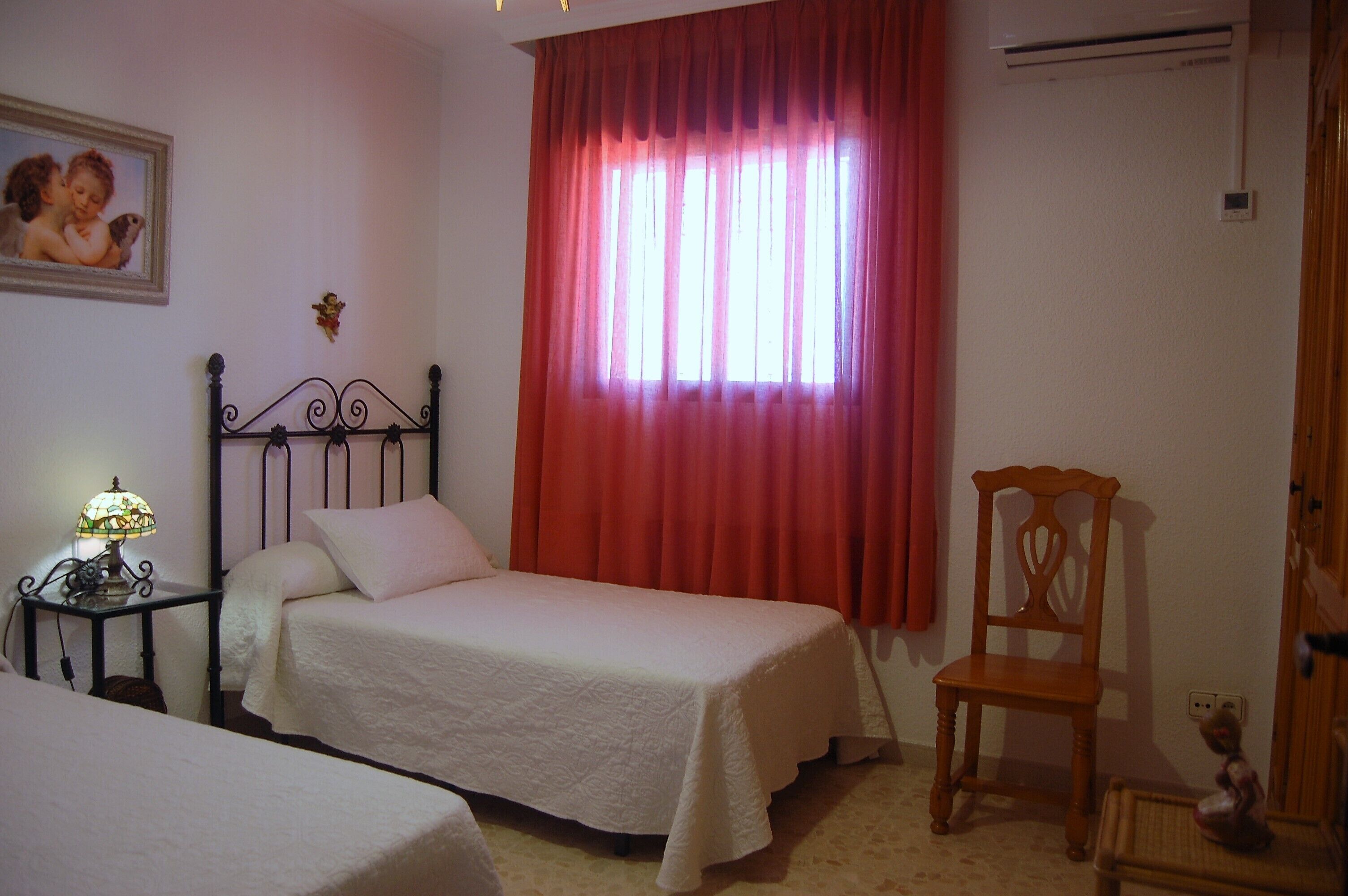 Une chambre avec deux lits simples, une table de chevet avec une lampe, une chaise en bois et une fenêtre avec des rideaux rouges.