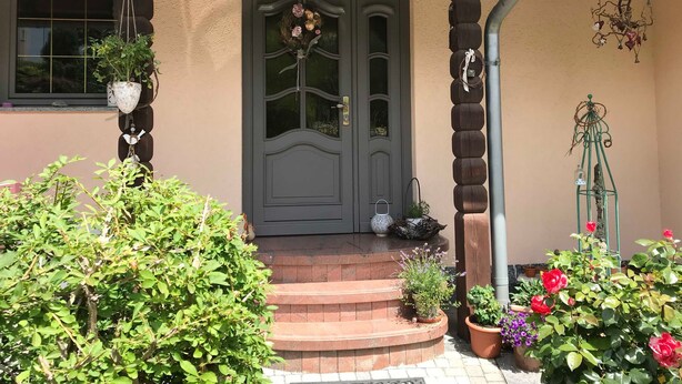 Une entrée de maison avec une porte de couleur gris foncé, un petit porche et diverses plantes en pot.