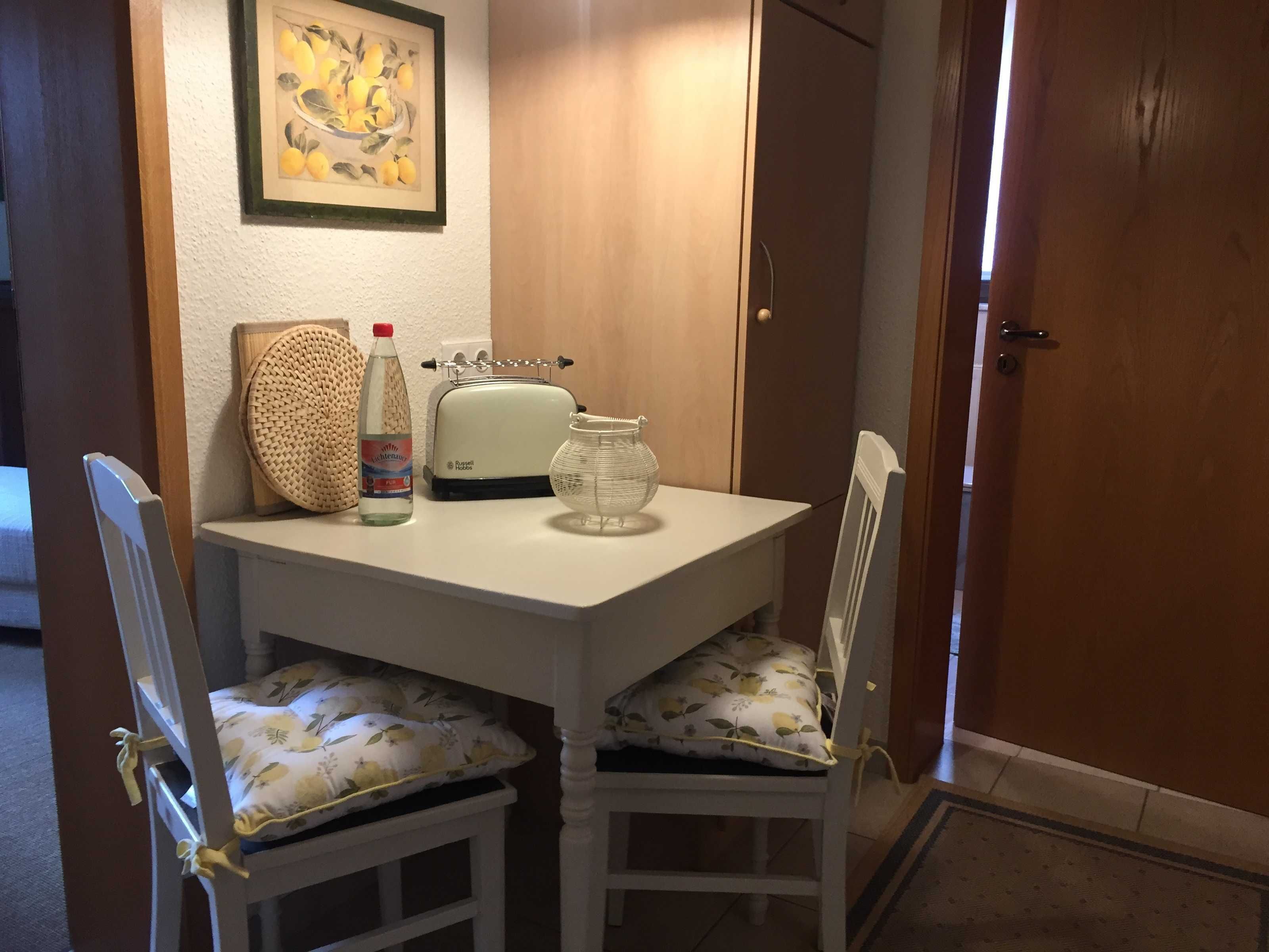 Ein kleiner Essbereich mit einem weißen Tisch, zwei Stühlen, einer Flasche, einem Toaster und einem gerahmten Bild an der Wand.