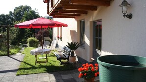 Une terrasse avec une table et des chaises, un parasol rouge, un banc et une poubelle.