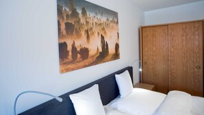 Una cama bien hecha con sábanas blancas, un armario de madera y un cuadro con una escena de bosque en la pared.
