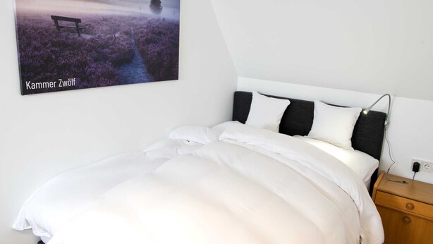 Une chambre avec un lit blanc, une table de chevet, un tableau accroché au mur et un banc placé dans un champ.