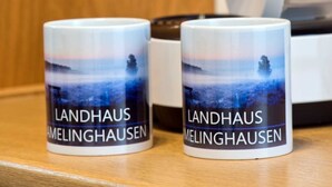 Två muggar med ett landskapsmotiv och texten ”Landhaus Melinghausen” tryckt på dem, placerade på en träyta.