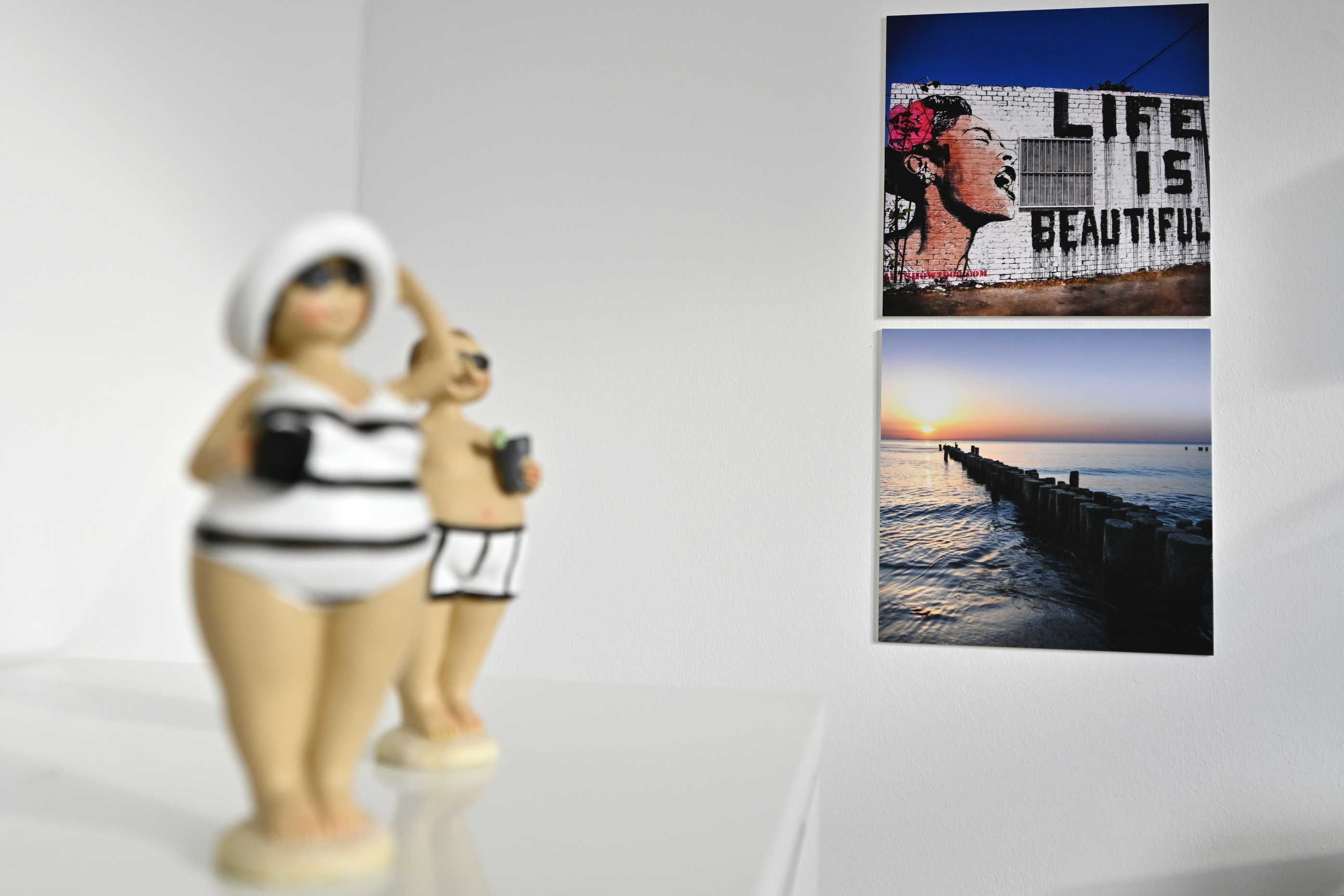 A room with a white wall, a small statue of a woman and a child, and two framed pictures depicting a sunset and a quote.