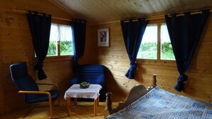 A cozy cabin room with wooden walls, a bed, a chair, a table with a decorative item, and curtains. - Frehmeyer: Vacation on the farm - Ferienhaus Frehmeyer (Lindewitt)