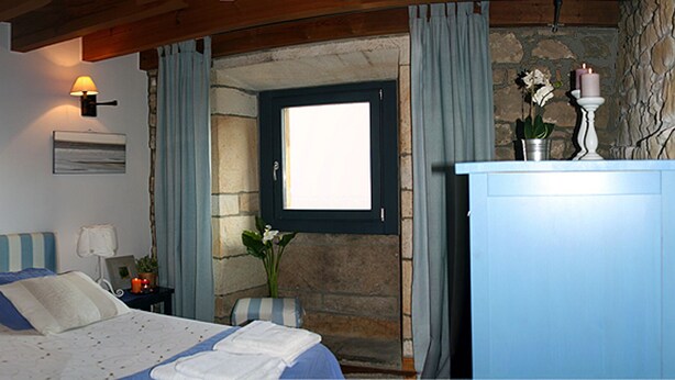 A bedroom with a bed, bedside lamps, a nightstand with a plant, a dresser, and a stone wall. - Alojamiento Rural con Encanto La Casona del Piquero (Lastres)