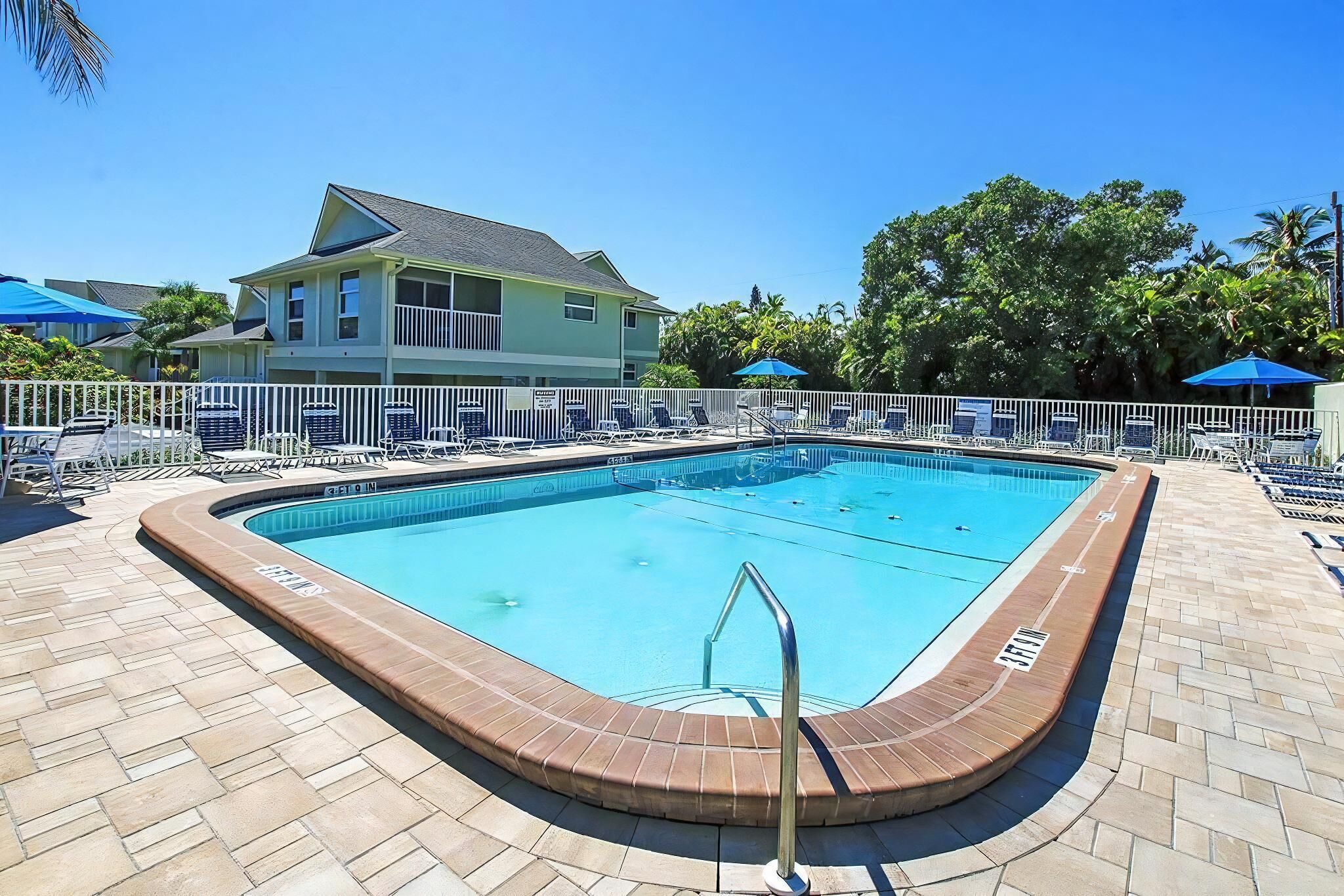 A swimming pool with a tiled deck, surrounded by lounge chairs and umbrellas, with a building in the background.