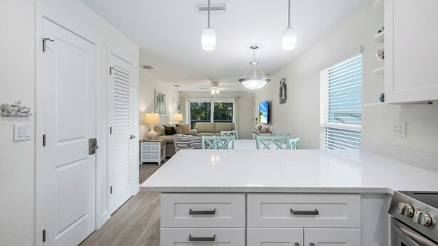 A modern kitchen with white cabinetry, a central island, and a dining area with teal chairs. - Seashells #39 |July & August Discounts! 3 Night Min. Stays! (Sanibel)