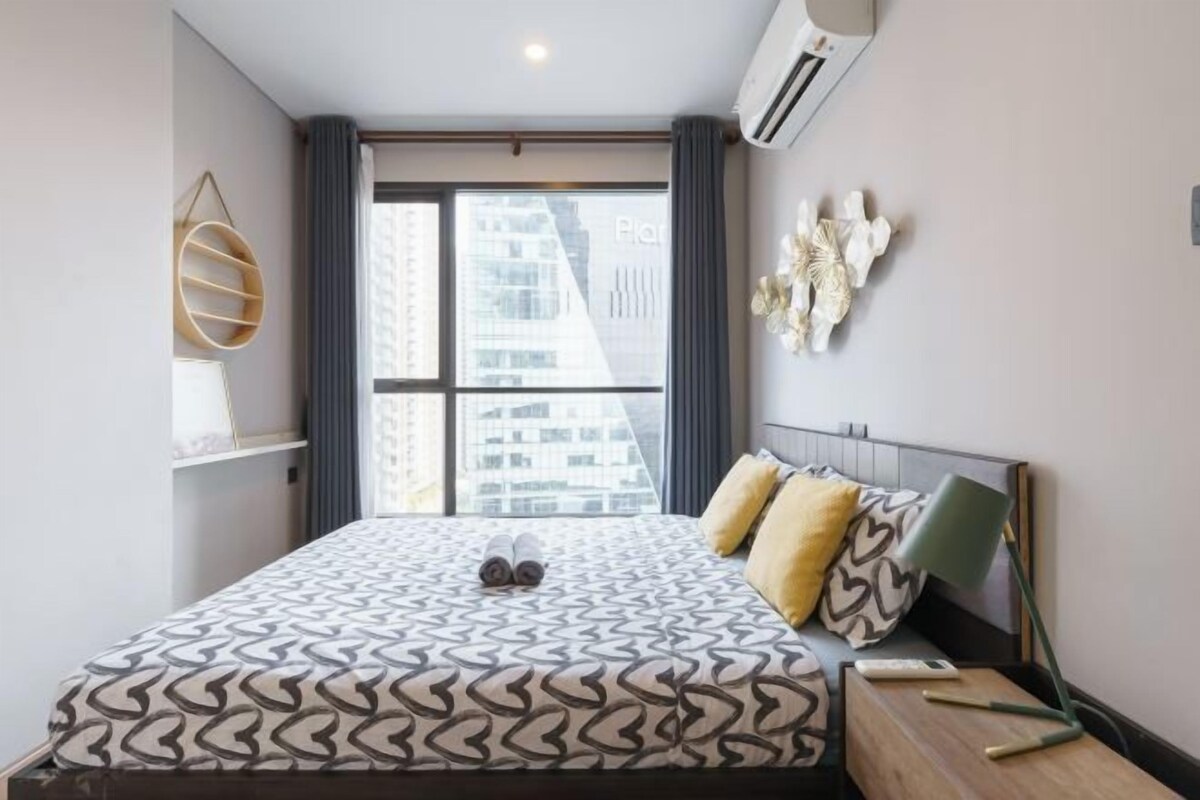 A modern hotel room with a large bed, a bedside table, a lamp, and a view of the cityscape through the window.