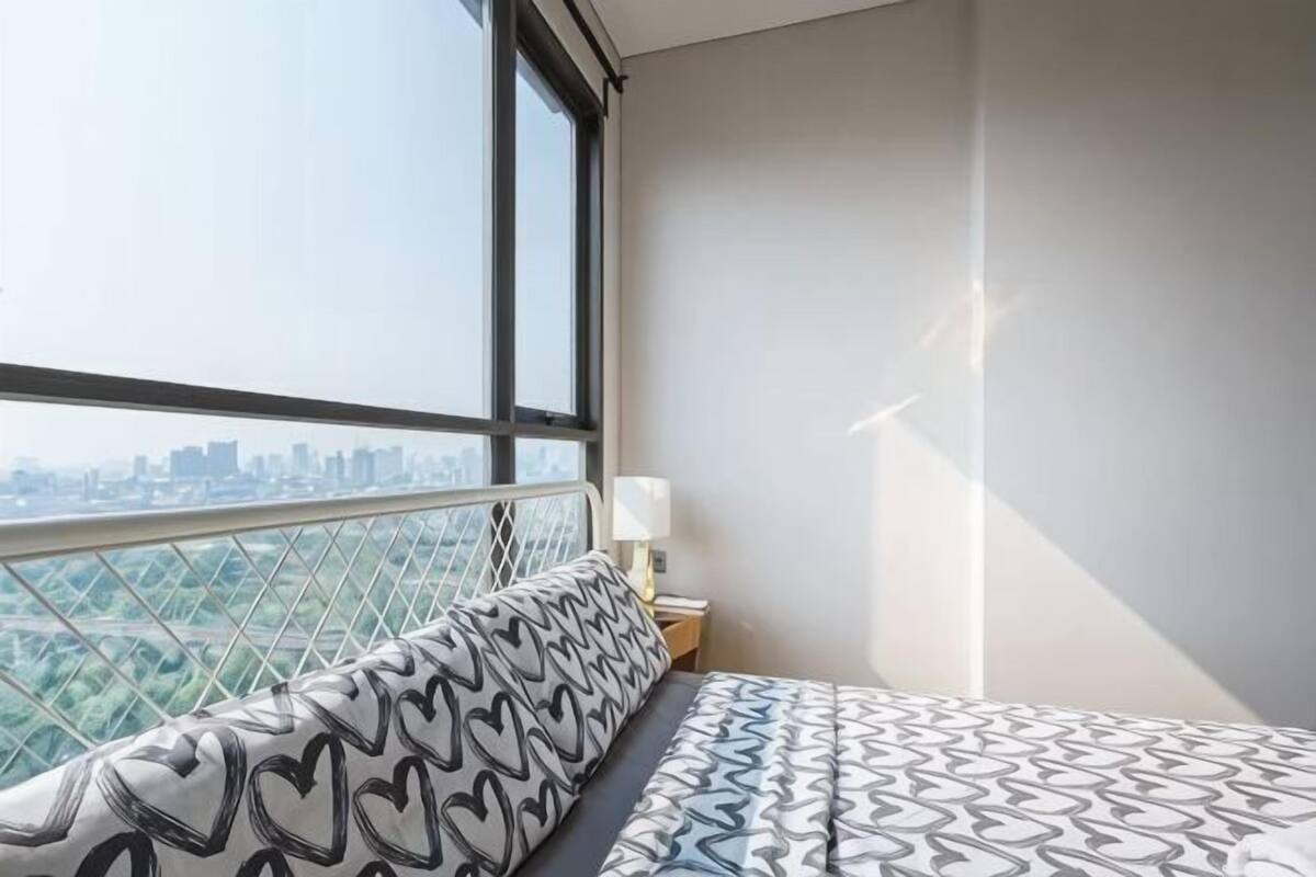 A bedroom with a city view, a patterned bedspread, a heart-patterned pillow, and a nightstand with a lamp.