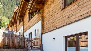 A wooden chalet with a patio area featuring a glass table and chairs, a wooden fence, and a view of a mountainous landscape. - Chalet Häuser (oetz)