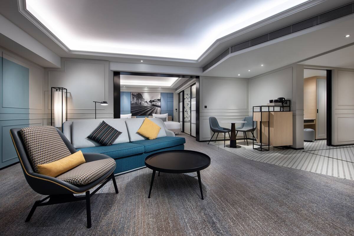 Un moderno vestíbulo de hotel con un sofá azul, un sillón estampado, una mesa de centro redonda y un área de comedor con una mesita y sillas.