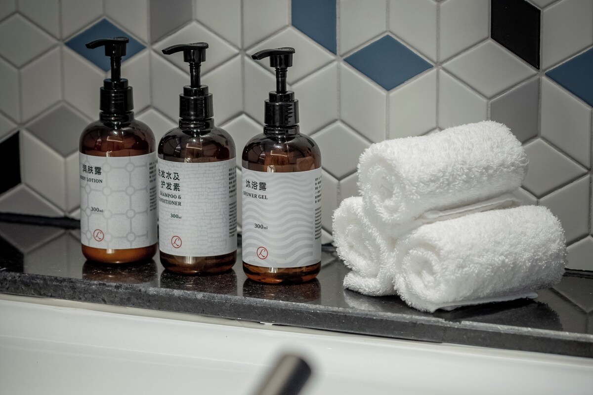 A bathroom with a tiled wall, three bottles of liquid soap, and a stack of white towels.