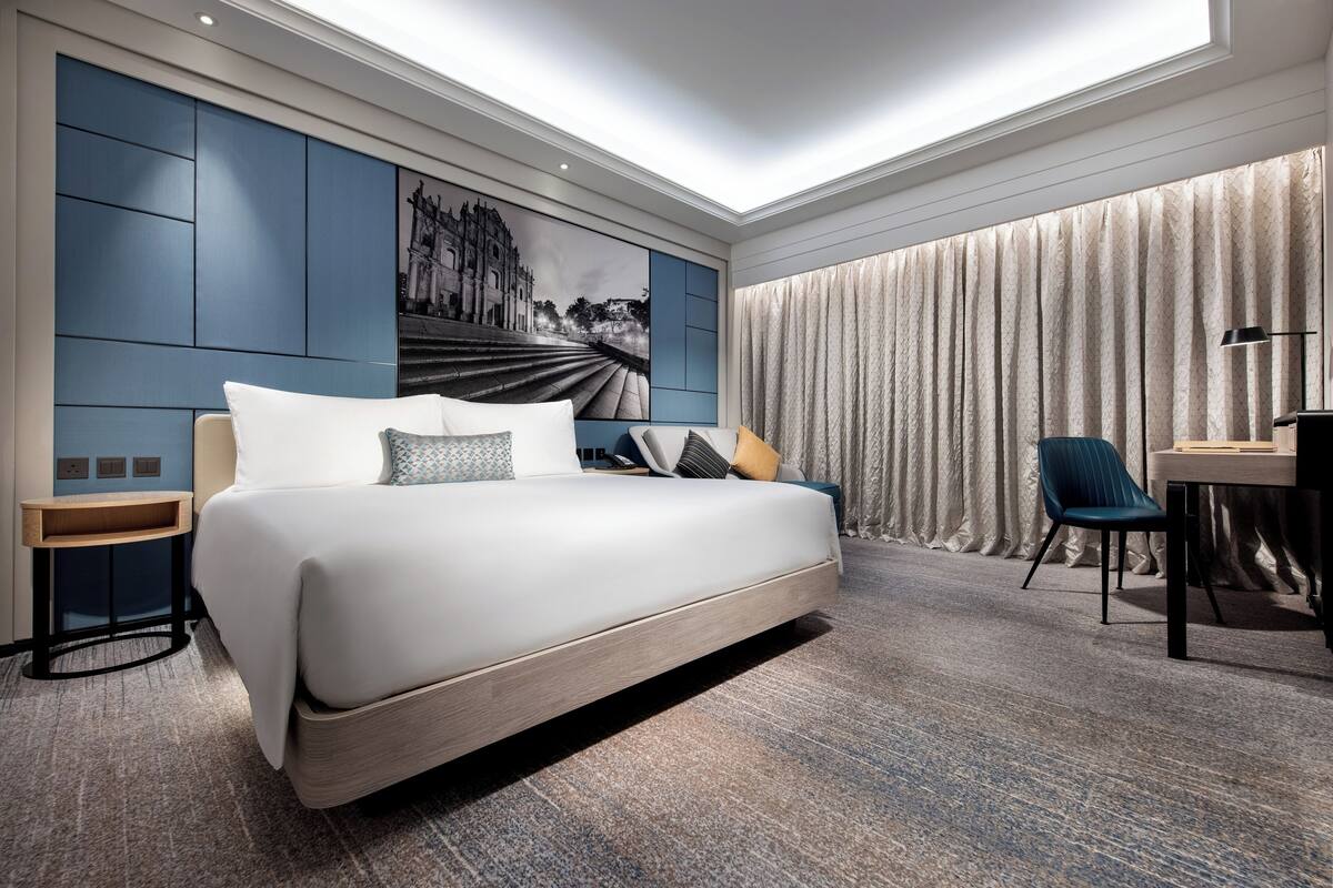 Una habitación de hotel moderna con una cama grande, un escritorio, una silla, un televisor montado en la pared y un amplio ventanal con cortinas.