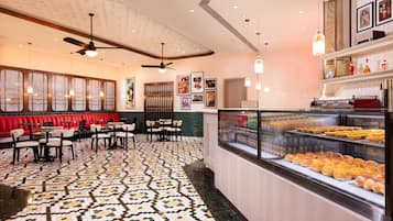 Un restaurante con piso estampado, zona de estar con mesas y sillas, y una vitrina con productos de panadería.