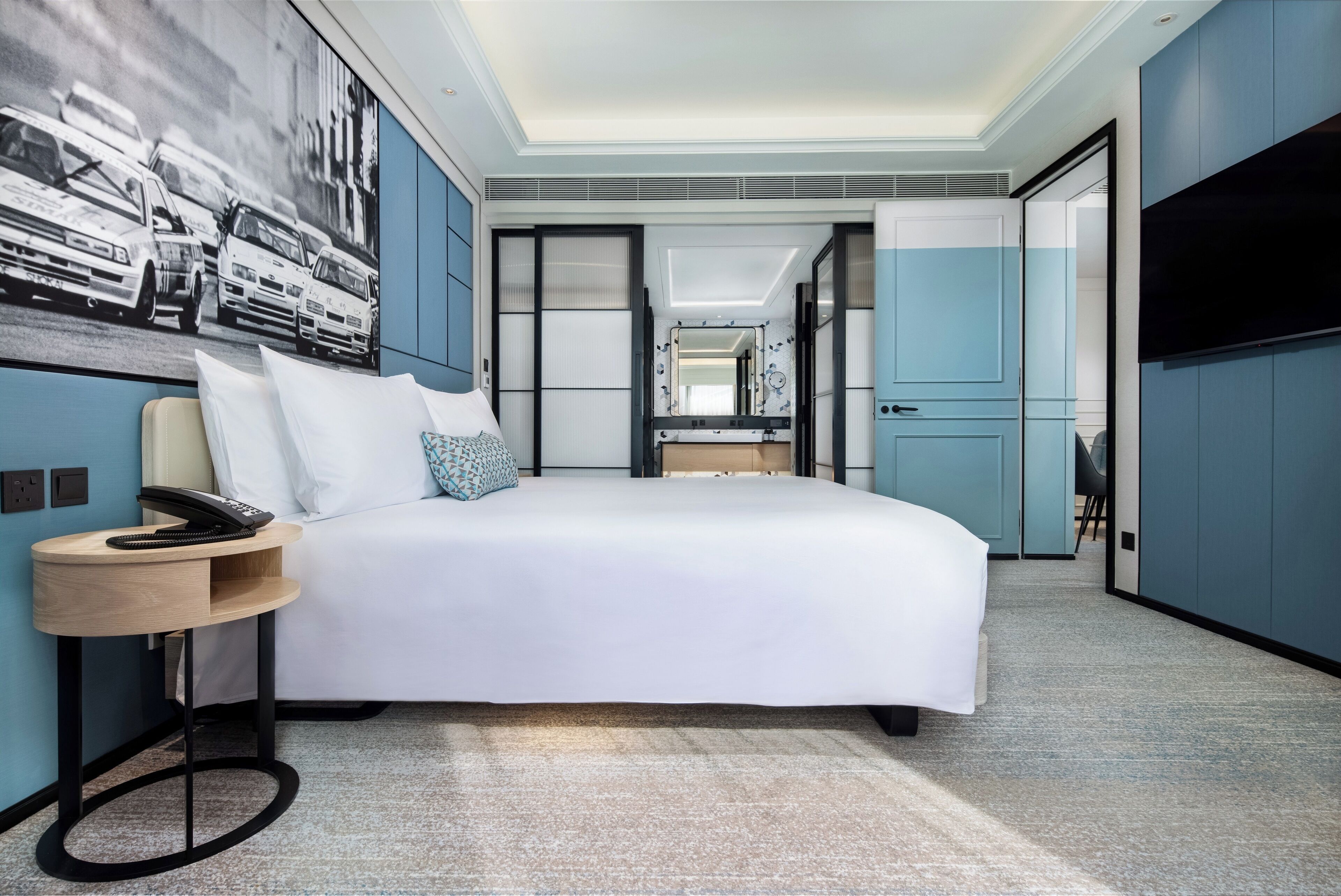 A modern hotel room with a large bed, a bedside table with a phone, a wall-mounted TV, and a painting of taxis.