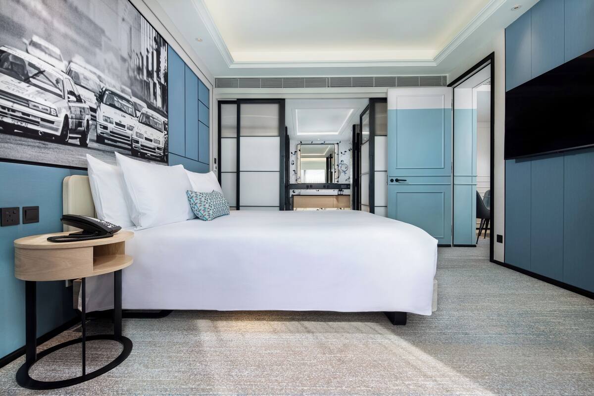 Una habitación de hotel moderna con una cama grande, una mesita de noche con teléfono, un televisor de pared y un cuadro con taxis.