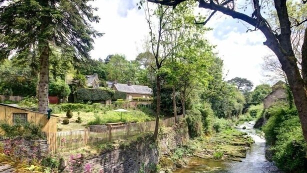 Een vredig landschap aan de rivier met een houten brug, weelderige begroeiing en een klein huis met rieten dak.