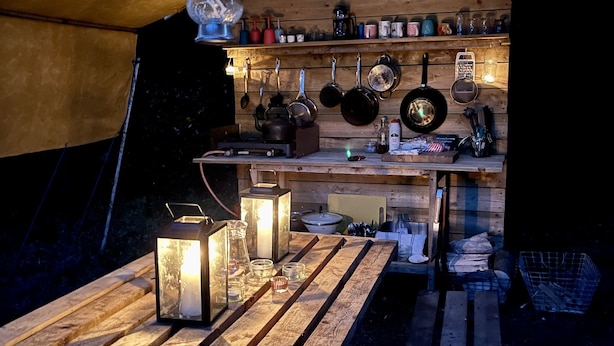Een rustieke keuken met houten planken, hangende pannen en een houten tafel met lantaarns en flessen.