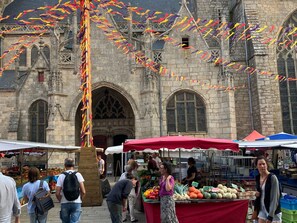 Un bâtiment historique à l’architecture gothique, un étal de marché proposant des produits frais, et des gens qui font leurs courses.