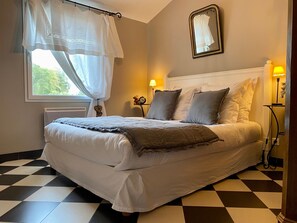 Ein ordentlich bezogenes Bett mit grau-weißer Decke, ein Nachttisch mit Lampe, einem Teddybären und einer Uhr.