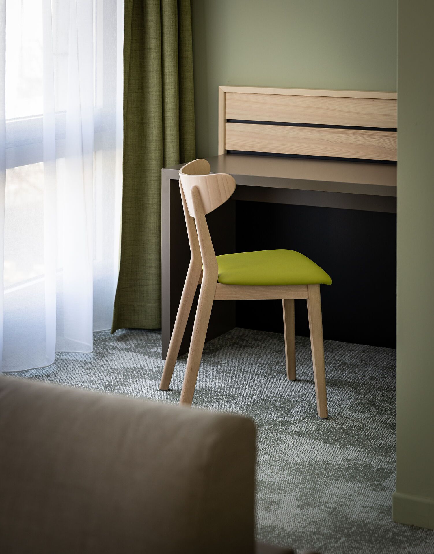 Een kamer met een houten stoel met een groene kussen, een bureau en een raam met dunne gordijnen.